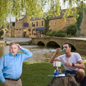 Saucepangate at Bourton-On-The-Water - a middle-aged man in a blue shirt is looking shocked at a younger man eating food from a saucepan on a camping stove